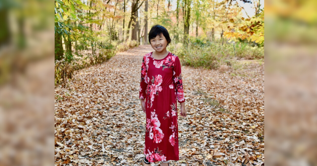 A young girl in a red dress stands amidst the trees in a serene forest setting.