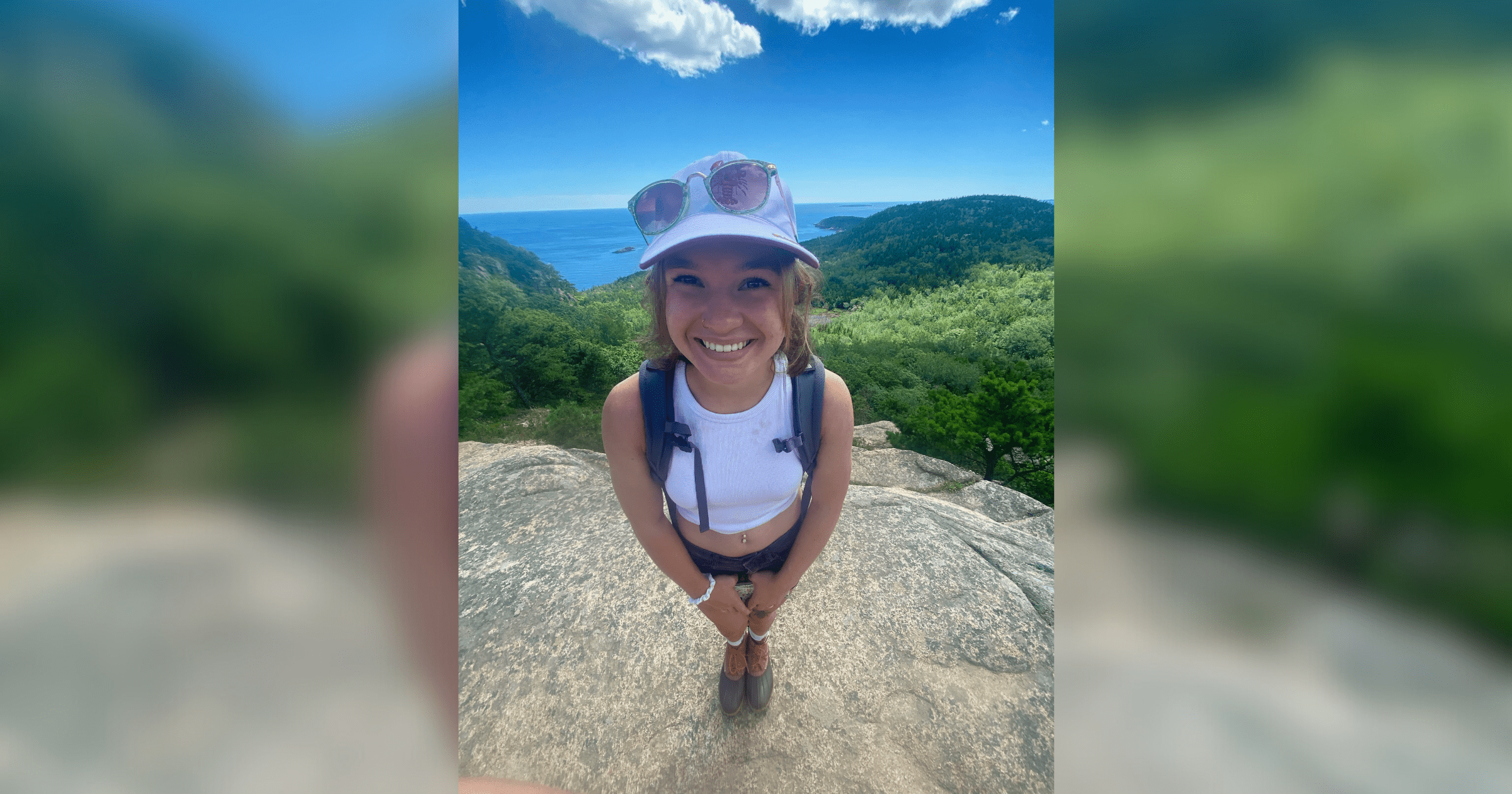 A woman wearing a hat and a white shirt stands confidently on a rock, enjoying the scenic view around her.