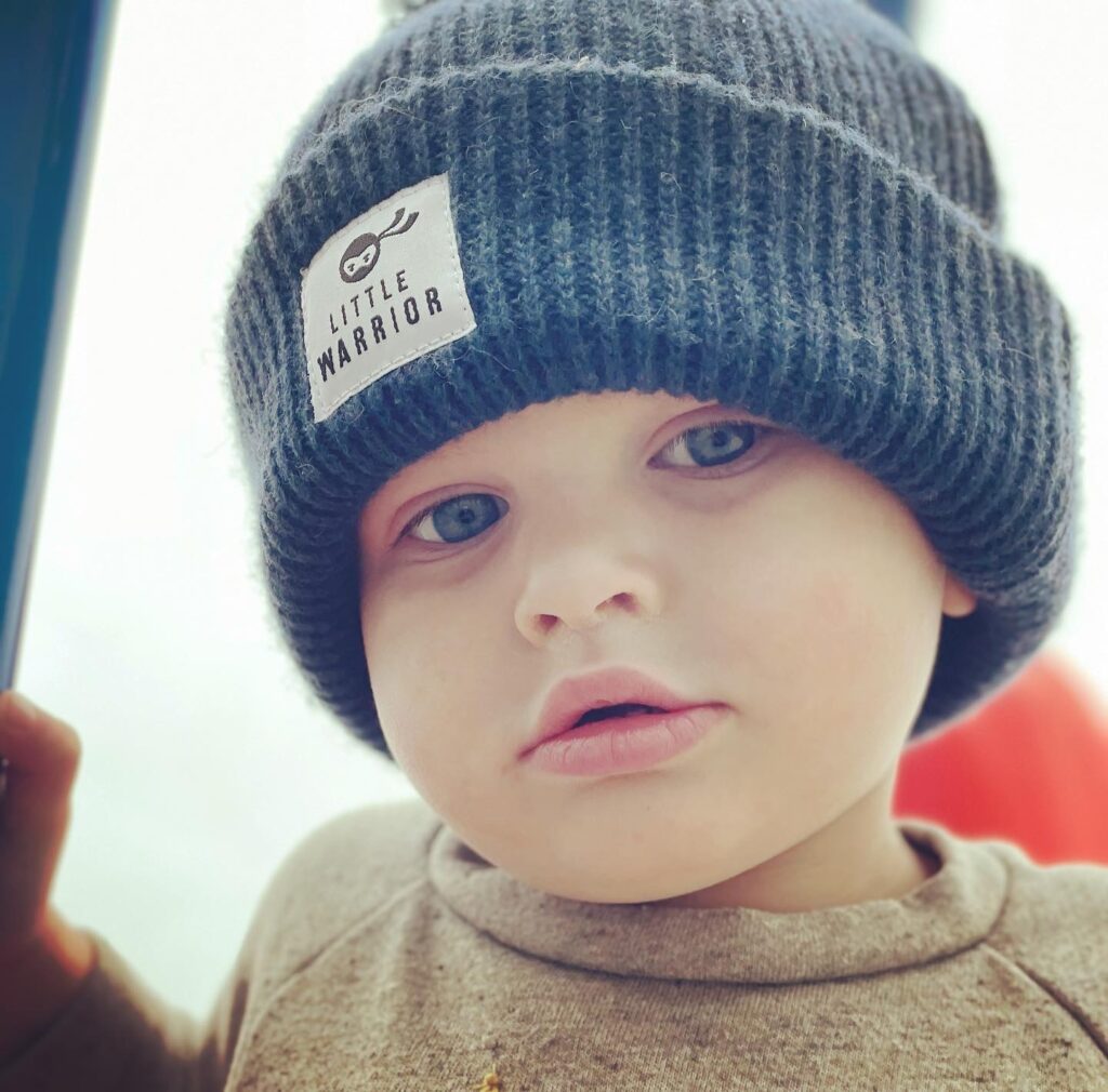 Little boy with cancer wearing a beanie that says, “little warrior.”