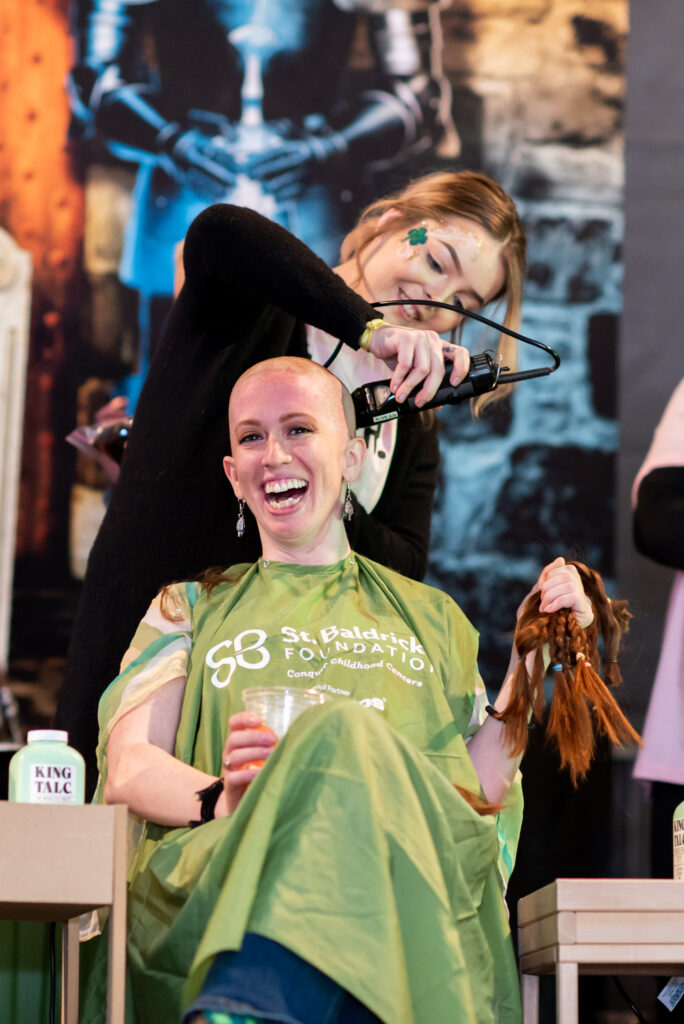 a female shavee who's all smiles, holding a drink in her right hand and her hair donation in the left