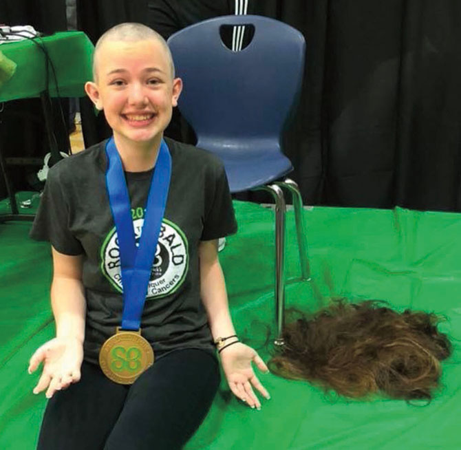 a female shavee with her new bald look with a medallion around her neck, while sitting next to a pile of her hair that was just shaved off