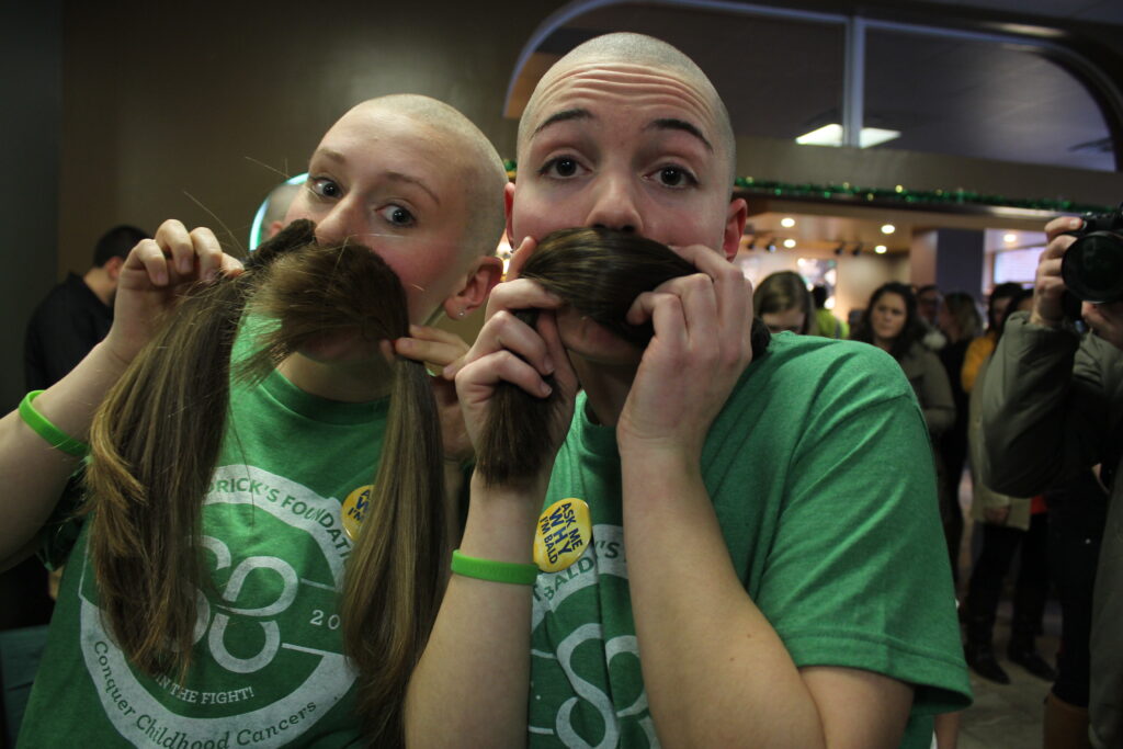 2 female shavees rocking the bald while holding their locks for hair donation as mustaches against their face