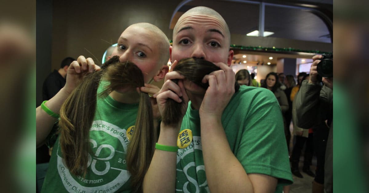 2 female shavees rocking the bald while holding their locks for hair donation as mustaches against their face