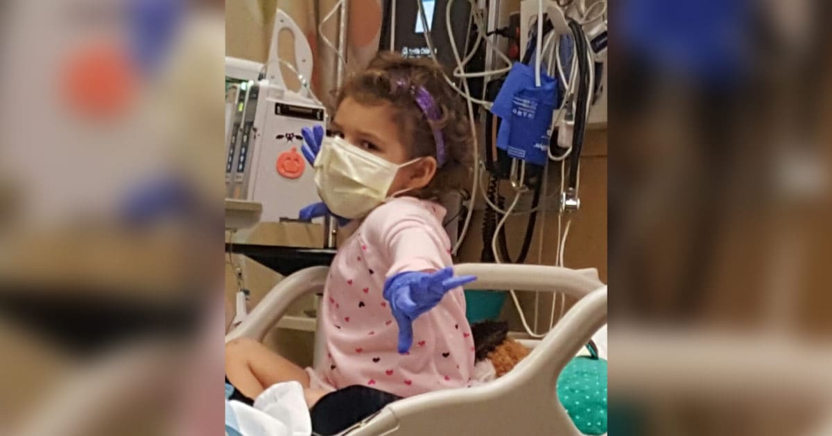 cancer warrior Massy in the hospital, wearing a purple headband, a pink shirt with tiny hearts, blue gloves, and a face mask