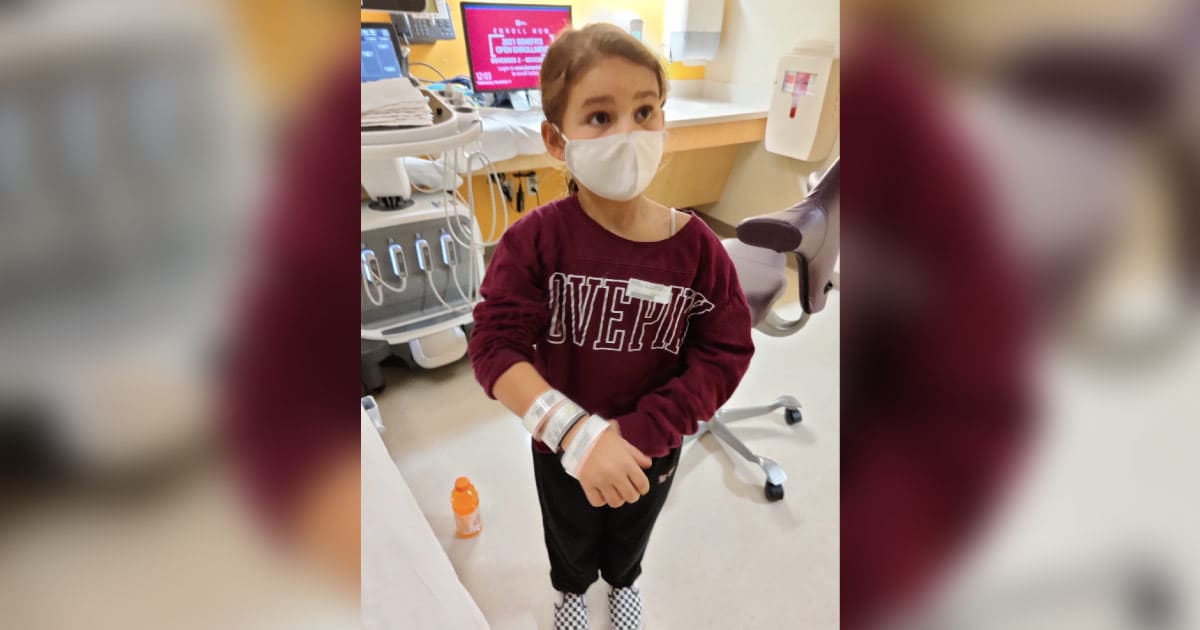 cancer warrior Massy in the hospital, wearing a maroon sweater with the words 'love pink' across the front, multiple hospital bands around her right wrist, black pants, black and white checkered shoes, and a face mask