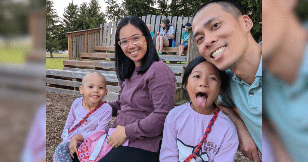 A family of four sits on wooden benches outdoors, smiling and making playful faces. It's a sunny day with green trees in the background.