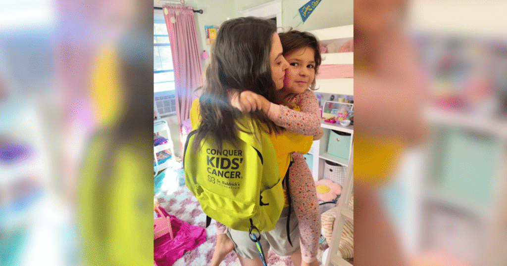 A woman wearing a bright green backpack with the words "Conquer Kids' Cancer" kisses a young child on the cheek in a cozy, colorful bedroom.