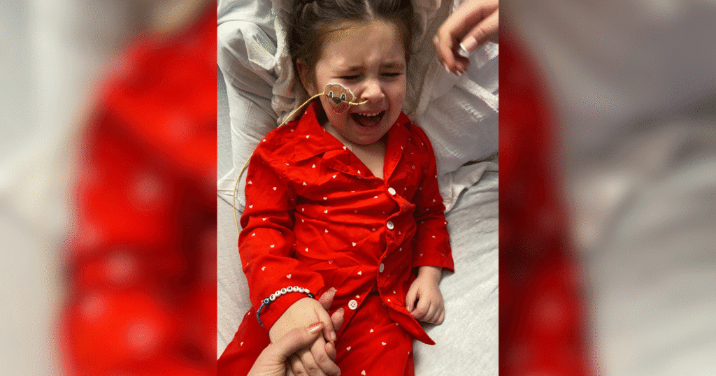 A crying girl in a red shirt with a nasal tube in the hospital.