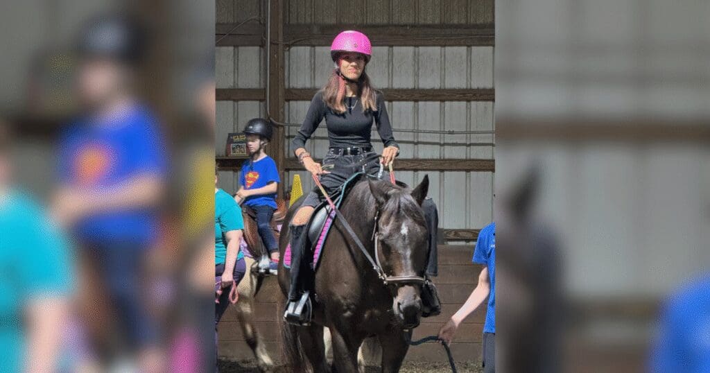 A woman wearing a pink helmet rides a black horse indoors. She smiles, flanked by people in blue shirts, creating a lively, safe atmosphere.