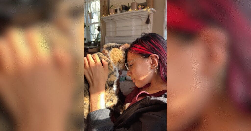 A person with red-tinted hair gently touches foreheads with a tabby cat, creating an intimate moment. Soft lighting filters in from a nearby window.
