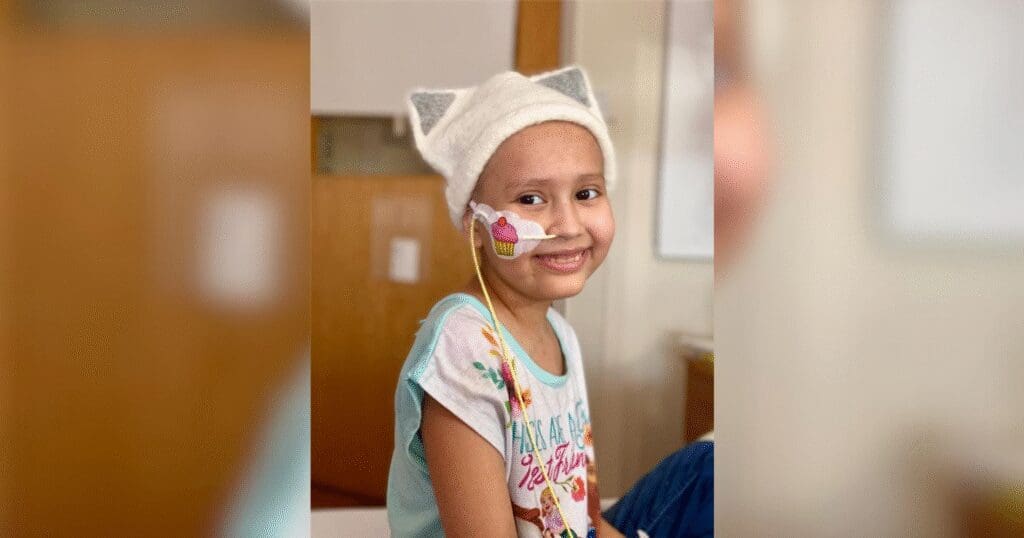 A smiling child wearing a cat-ear hat and a colorful shirt sits in a hospital setting. A nasal tube is visible, conveying resilience and warmth.