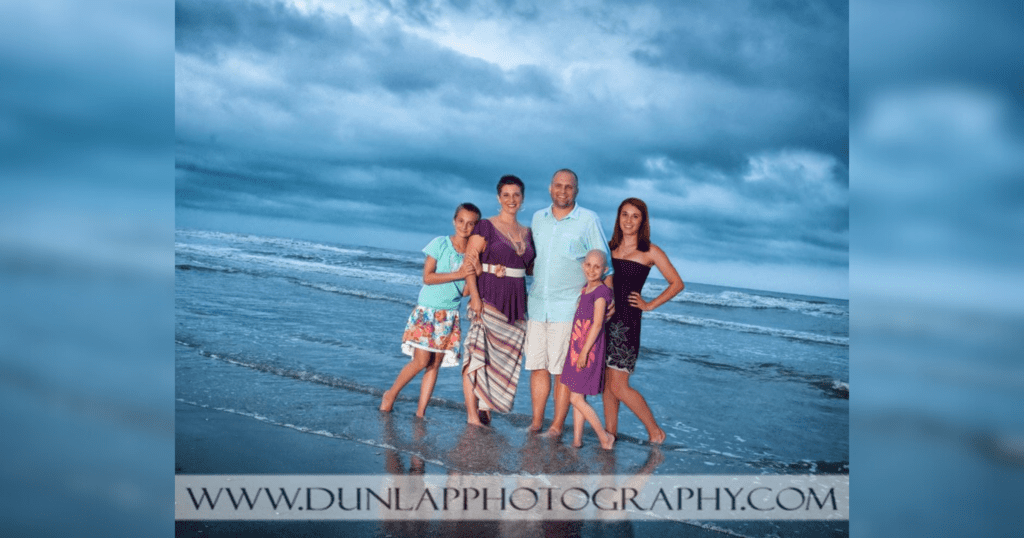 A family stands together on the beach, smiling for a portrait with the ocean waves in the background.