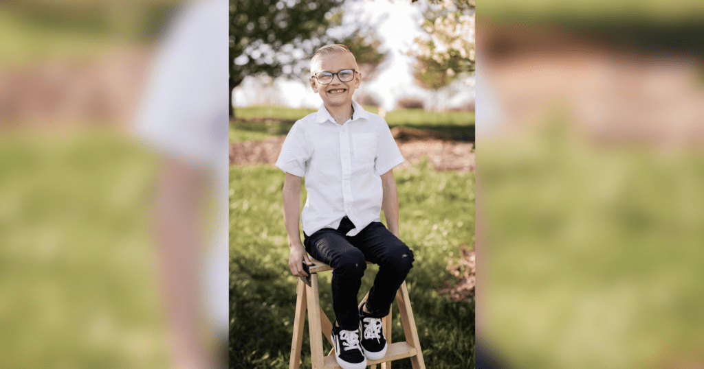 A young boy with glasses is seated on a chair, all smiles.