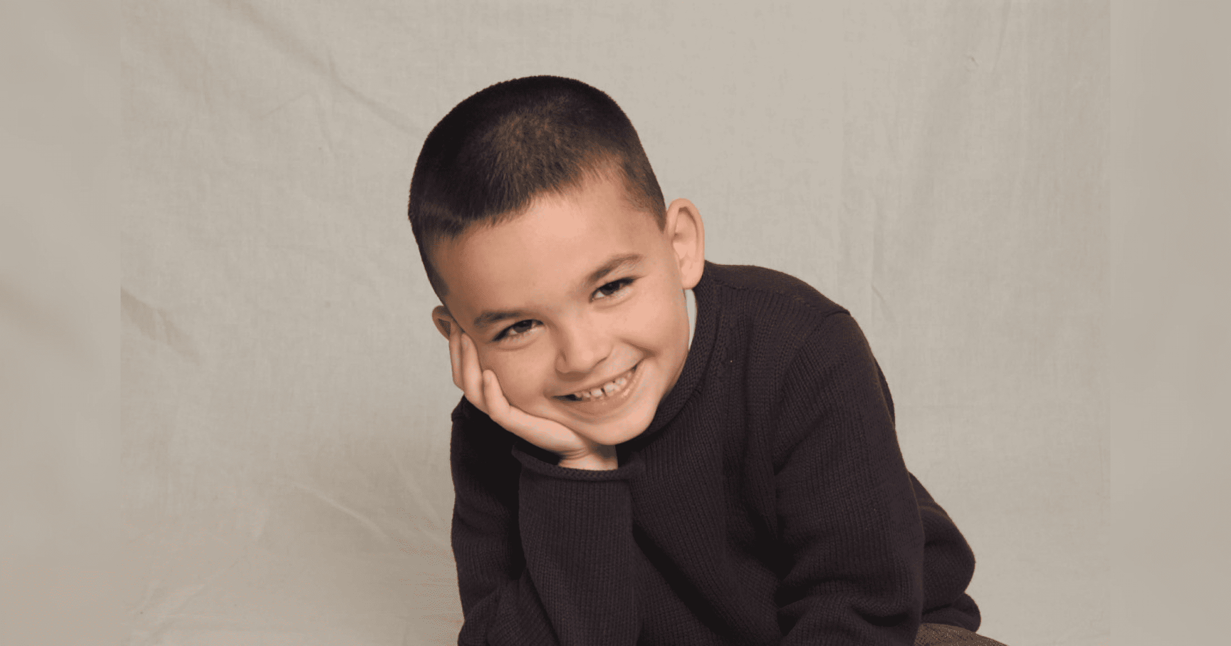 A young boy smiles brightly with his hand on his cheek, exuding joy and playfulness in a relaxed position.