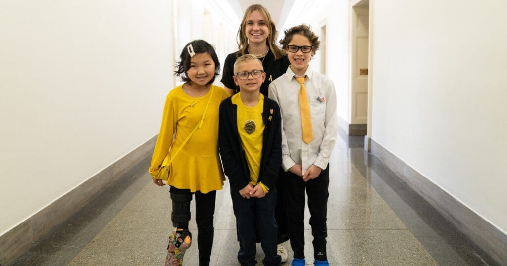 A joyful group of children and a woman stands in a bright hallway. The children wear matching yellow-themed outfits, smiling warmly at the camera.