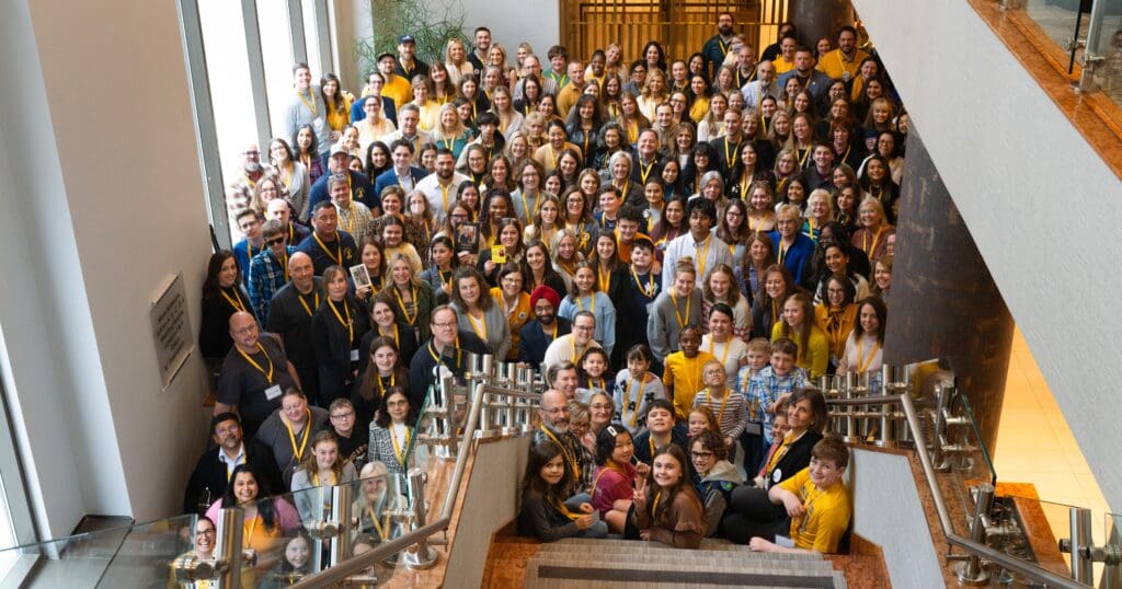A large group of people, diverse in age and appearance, smile and pose on a staircase in a spacious lobby. Many wear yellow lanyards, creating a unified and cheerful atmosphere.