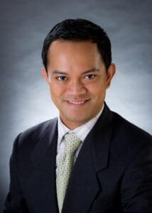 Smiling man in a suit and tie with a light gray background. He projects a professional and approachable demeanor.