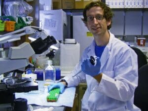 A scientist in a white lab coat and gloves smiles while holding a pipette in a lab. He's surrounded by microscopes, test tubes, and lab equipment, creating a focused yet friendly atmosphere.