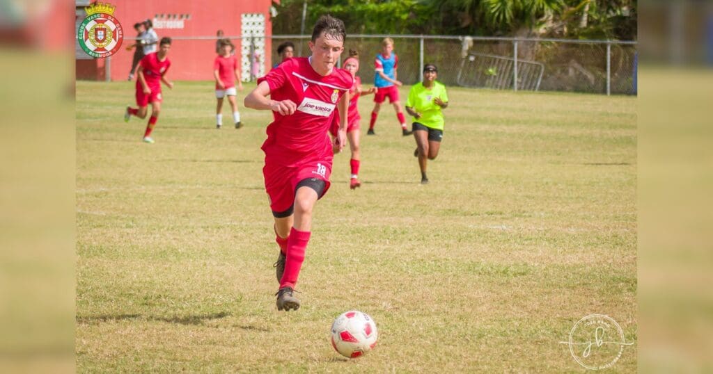 A soccer player in a red uniform, focused and determined, runs towards the ball on a grassy field. Teammates and a referee are visible in the background.