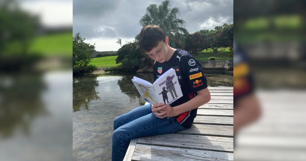 Teenage boy sits on a wooden dock reading a book, surrounded by tranquil water and lush greenery. He wears a racing-themed shirt, appearing focused and relaxed.