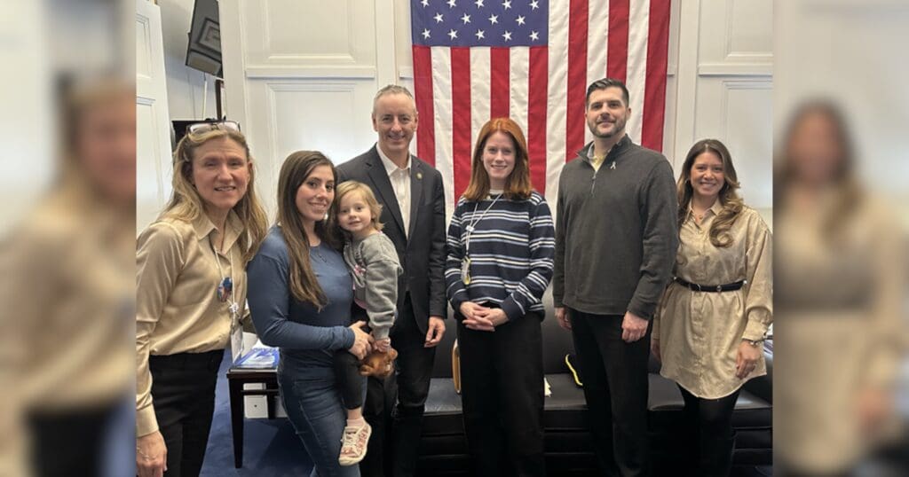 A group of six adults and one child stand smiling in a room with an American flag. The atmosphere is formal yet friendly.