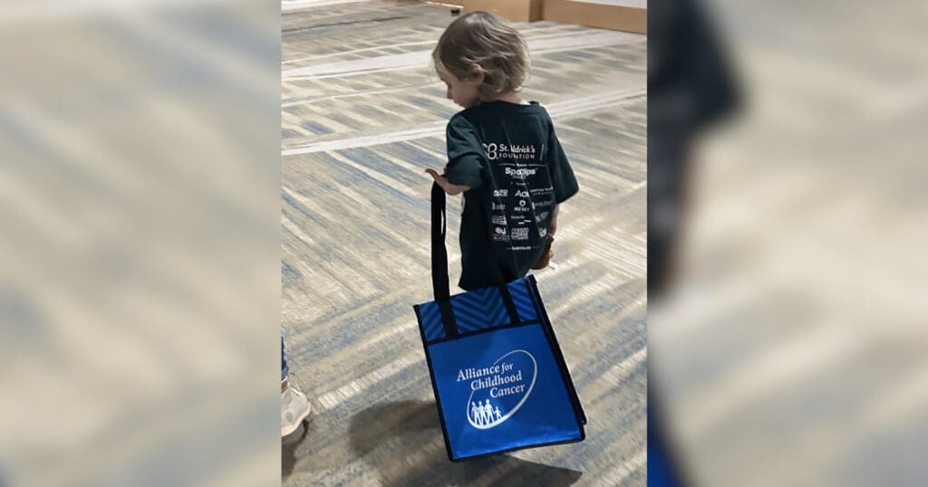 A young child with short hair walks on a patterned floor, holding a blue tote bag labeled "Alliance for Childhood Cancer." The scene feels hopeful.