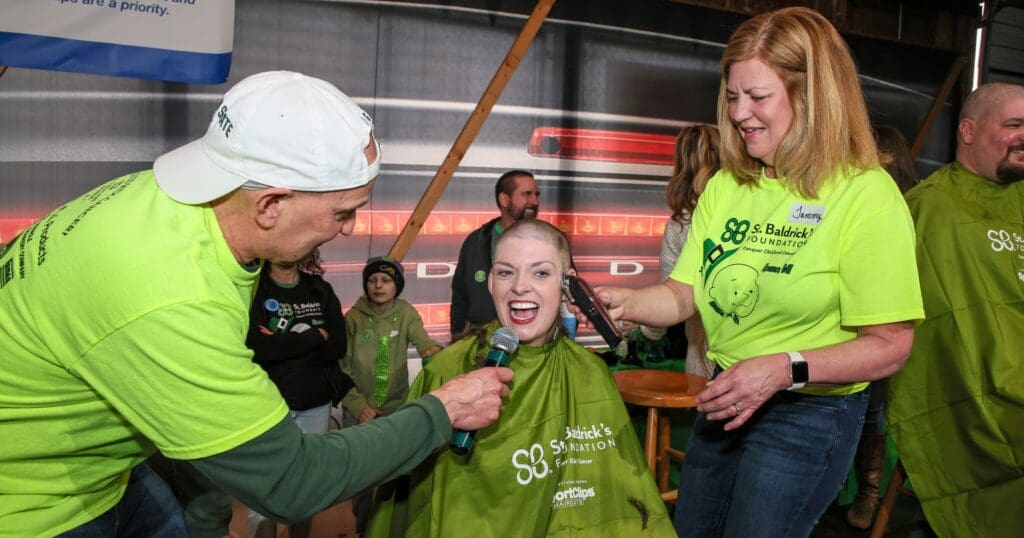 A woman smiles brightly while getting her head shaved at a St. Baldrick's Foundation event. She is covered with a green cape, surrounded by volunteers in matching shirts.