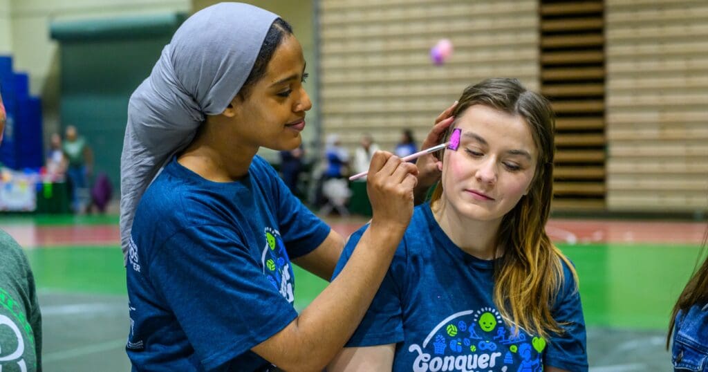 Two women in matching blue event shirts participate in face painting. One paints colorful designs on the other's face in a lively indoor setting.