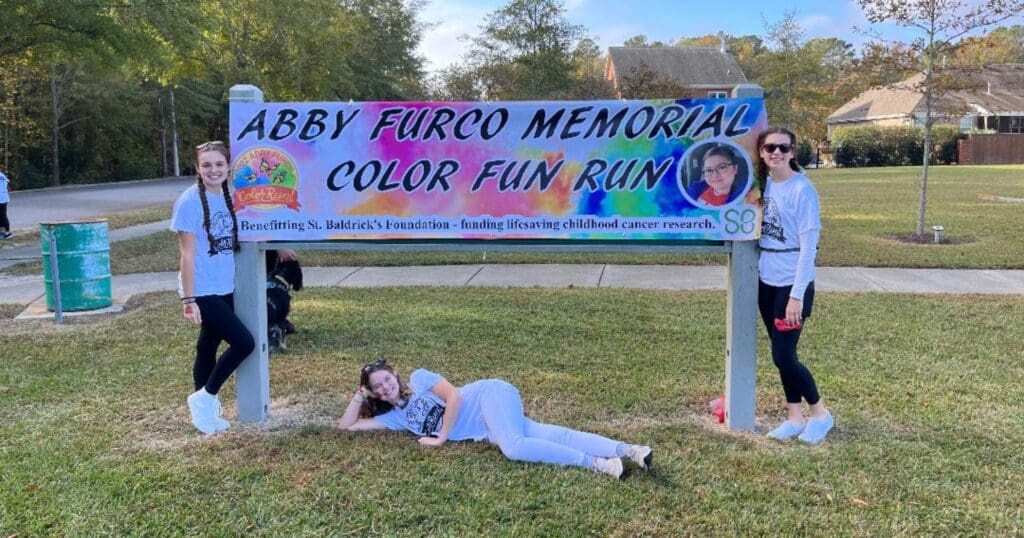 Three people pose happily by a colorful outdoor sign for the "Abby Furco Memorial Color Fun Run," set against a backdrop of trees and a suburban area.