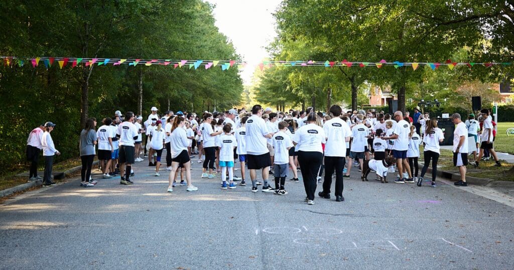 A large group of people in white shirts gather on a tree-lined road for a community fun run. Colorful flags are strung overhead, creating a festive atmosphere.