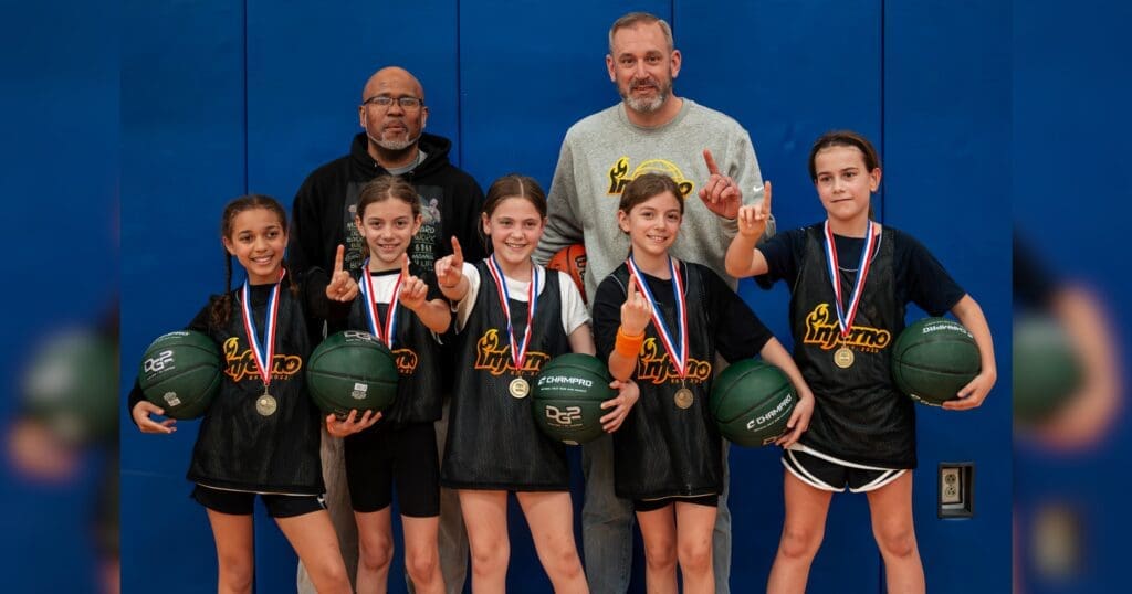 A basketball team of five young girls in black jerseys, wearing medals, smiles confidently with two coaches. They hold balls and gesture "number one."