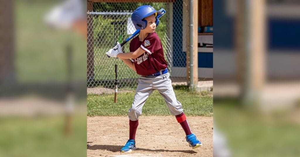Young baseball player in maroon jersey and blue helmet poised to swing, standing in a sandy field, with a determined expression. Chain-link fence in background.