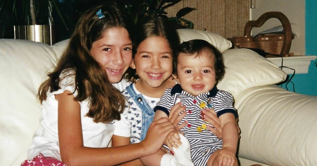 Three smiling children sit closely on a white couch. Two girls are on either side, hugging a baby boy in a striped outfit. The mood is joyful and warm.