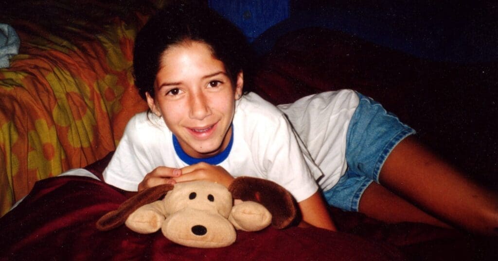 Girl with a bright smile lying on a bed, holding a stuffed dog toy. She wears a white top and denim shorts. The setting is warm and cozy.
