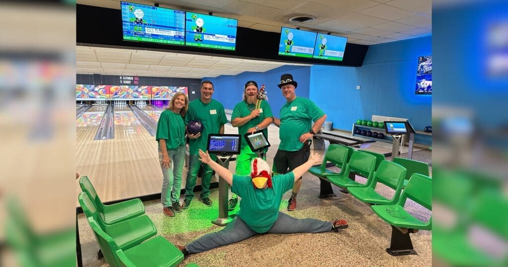 Five people in green shirts at a bowling alley, four standing with bowling balls and one doing a split wearing a chicken hat, conveying fun and excitement.