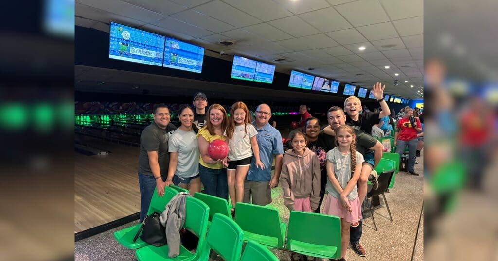 A group of smiling people pose in a bowling alley. A girl holds a pink bowling ball. The background displays bowling lanes and scoreboard screens.
