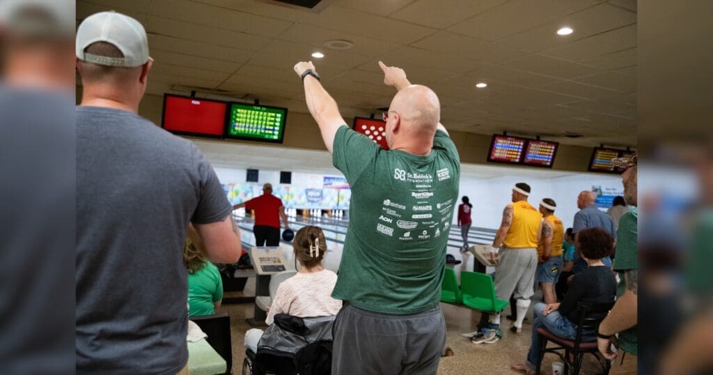 A lively bowling alley scene with people watching a game. A bald man in a green shirt enthusiastically raises his arms, celebrating a successful play.