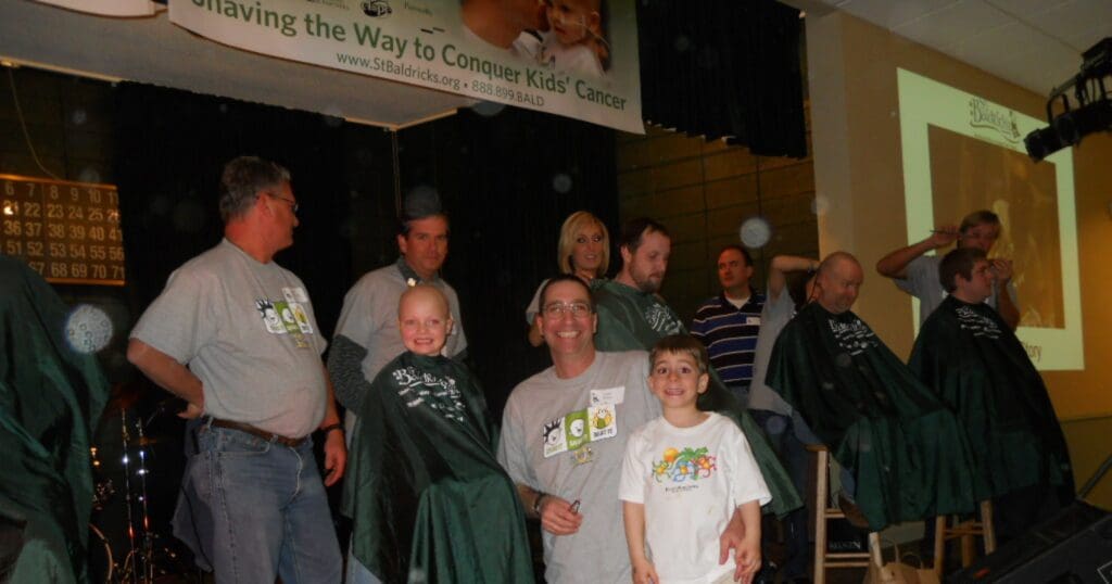 Group of people at a charity head-shaving event for children's cancer, smiling with camaraderie. Participants have shaved heads, creating a supportive, uplifting atmosphere.
