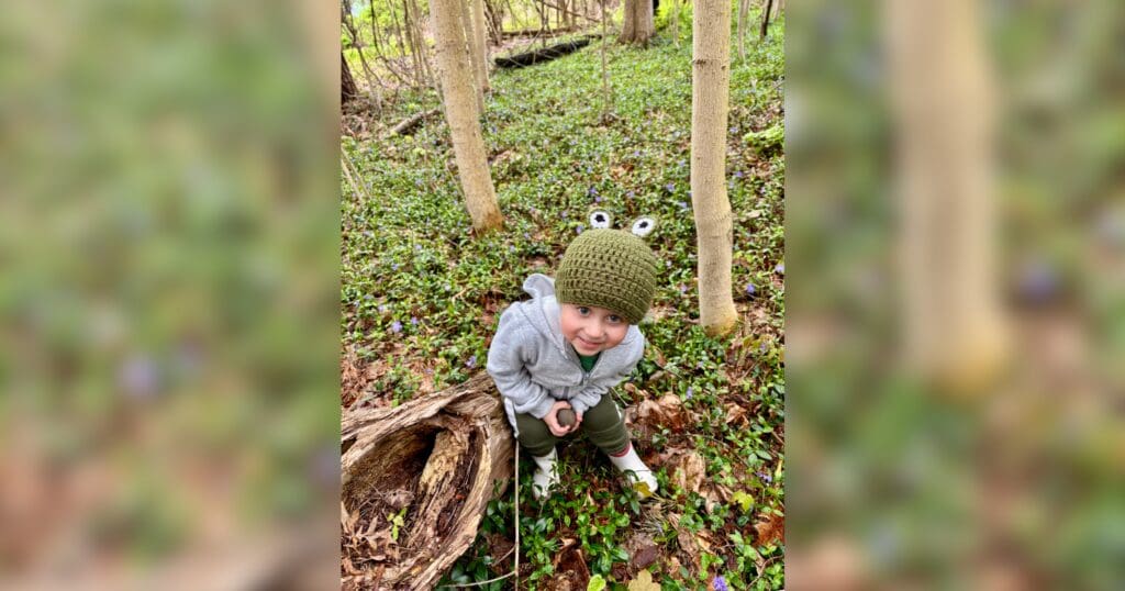A young child wearing a green frog hat crouches beside a fallen log in a wooded area covered with green ground plants, smiling at the camera.