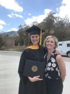 A woman in a graduation cap and gown stands beside another woman, both smiling proudly.