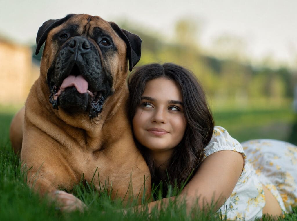 Young woman in a floral dress cuddles a large brown mastiff in the grass, both gazing at the camera with a warm, affectionate mood.