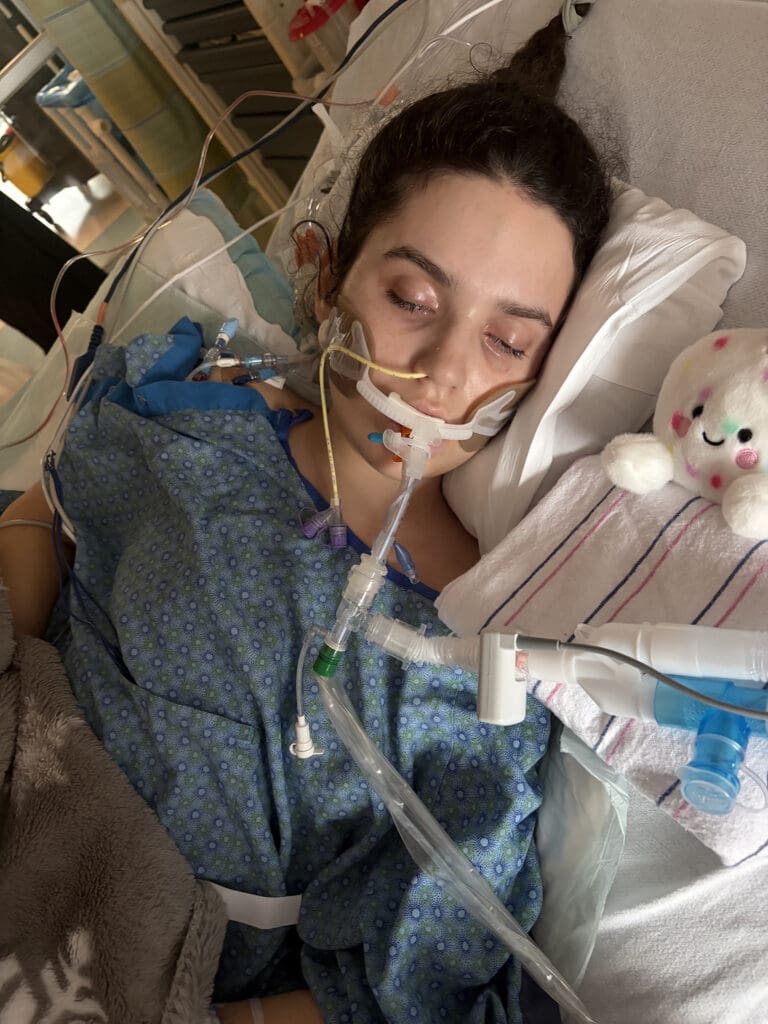 Hospitalized woman rests in bed with an oxygen tube, IV lines, and a small teddy bear beside him, conveying vulnerability and a somber mood.
