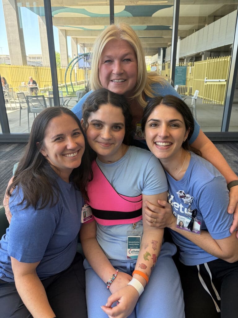 Four smiling women pose closely together indoors, wearing casual clothes and event badges, conveying friendship, warmth, and camaraderie.