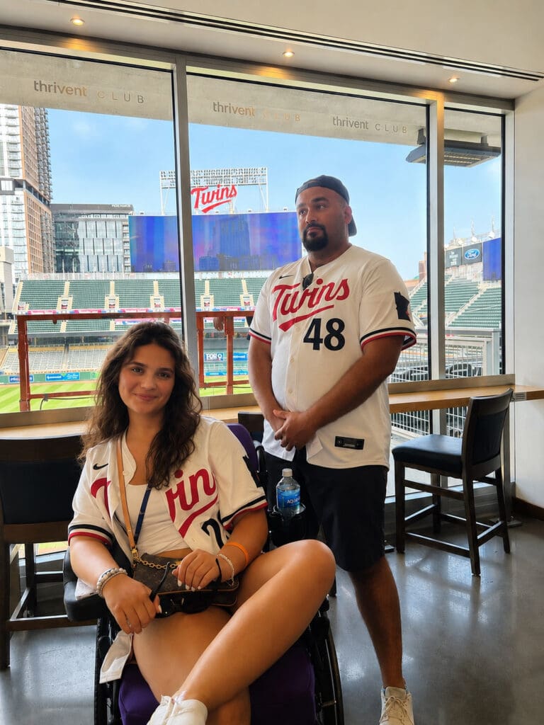 Two smiling Minnesota Twins fans pose in team jerseys inside a stadium restaurant, with Target Field and the baseball field visible through the windows.
