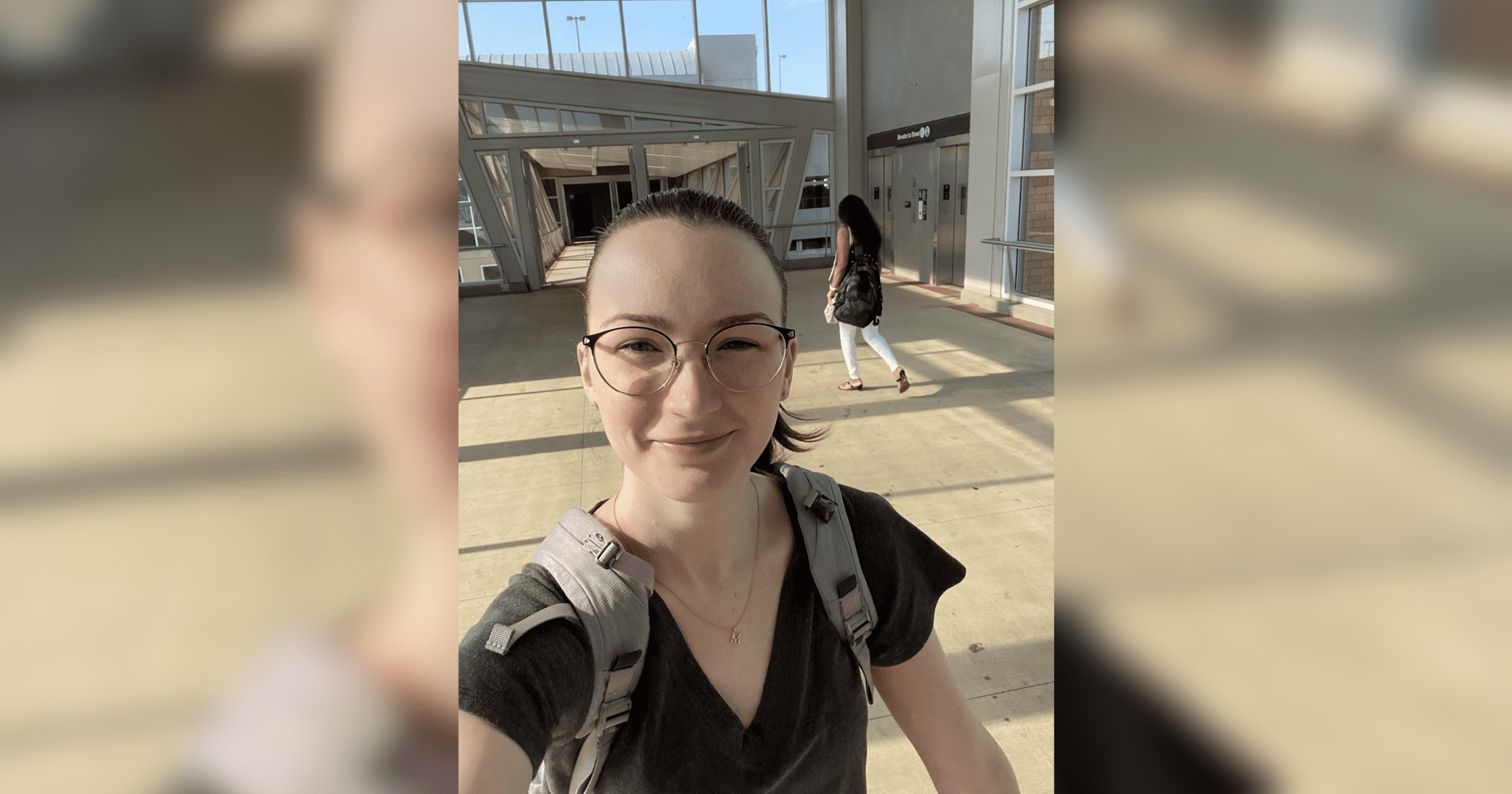 A woman with glasses and a backpack smiles while taking a selfie outdoors.