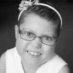 Black-and-white portrait of a smiling young girl wearing glasses and a bow headband, facing the camera against a dark background; cheerful mood.