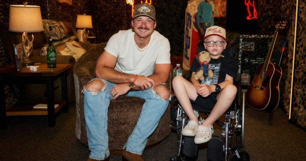 A man in a cap and ripped jeans sits smiling beside a child in a wheelchair holding a stuffed toy. A guitar and lamps add a cozy atmosphere.