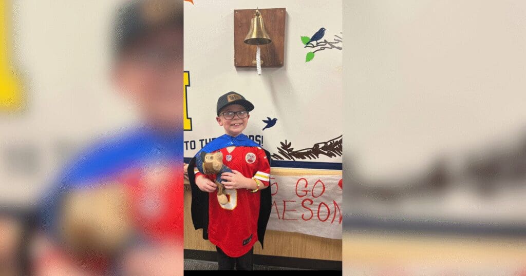 A young boy wearing glasses, a red shirt, and a cape stands smiling in front of a bell on the wall. He holds a stuffed toy, conveying joy and celebration.