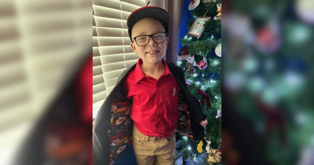 Young boy smiling, wearing glasses, a red shirt, cap, and opening a jacket lined with sports logos. Stands by a decorated Christmas tree. Joyful holiday vibe.