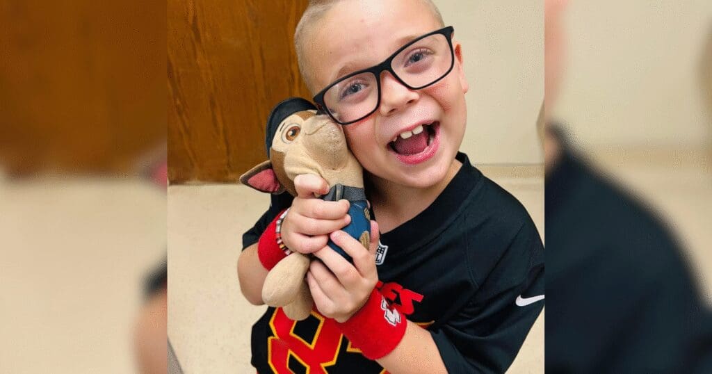 A young child with glasses smiles widely, hugging a stuffed toy. The child wears a black sports jersey and red wristbands, conveying joy and excitement.
